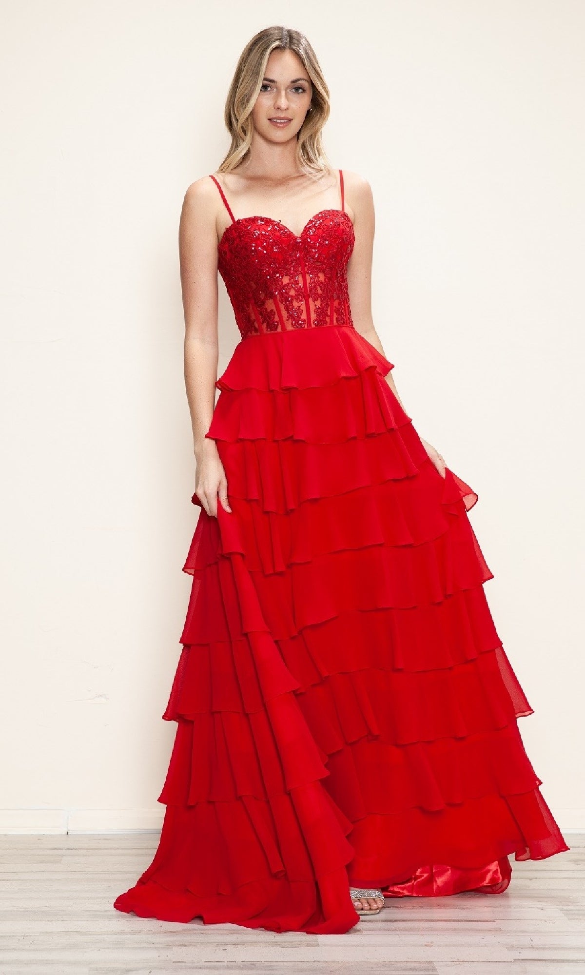 A woman in the Poly USA 9698 Long Tiered Corset Prom Dress, featuring a red a-line silhouette with a sequin-embellished fitted bodice and thin straps, stands against a plain light background.